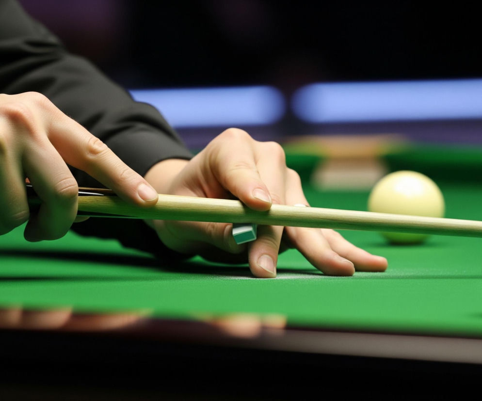 Lewa ręka osoby w czarnym rękawie koszuli tworzy mostek na zielonym suknie stołu bilardowego, podpierając jasnobrązowy kij. Prawa dłoń znajduje się dalej w tle, również trzymając kij. Na stole leży jedna biała i dwie czerwone bile, przy czym jedna czerwona kula z białym symbolem jest najbliżej prawej krawędzi kadru. Tło jest rozmyte, ukazując ciemne, niewyraźne kształty.