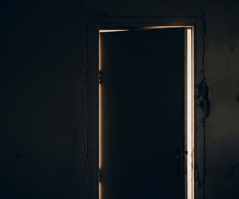 W centrum kadru znajduje się ciemne, lekko uchylone drzwi, otoczone z trzech stron jasną, pionową smugą światła wydobywającą się z wnętrza. Lewa strona zdjęcia oraz fragmenty ściany wokół futryny są pogrążone w głębokim cieniu, ukazując nierówną, pękniętą fakturę tynku. Na samej futrynie drzwi widoczny jest ciemny uchwyt.