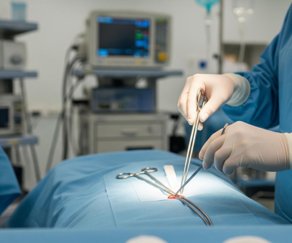 Na pierwszym planie widoczne są ręce w białych rękawiczkach chirurgicznych, trzymające metalowe narzędzia medyczne nad częścią ciała pacjenta, przykrytą niebieską tkaniną. Obszar roboczy jest jasno oświetlony reflektorem, ukazując fragment skóry z czerwonym szwem oraz inne narzędzia leżące obok. W tle znajdują się rozmyte urządzenia medyczne z ekranami wyświetlającymi dane i liczne kable, wszystko utrzymane w sterylnej, jasnej tonacji typowej dla sali operacyjnej.