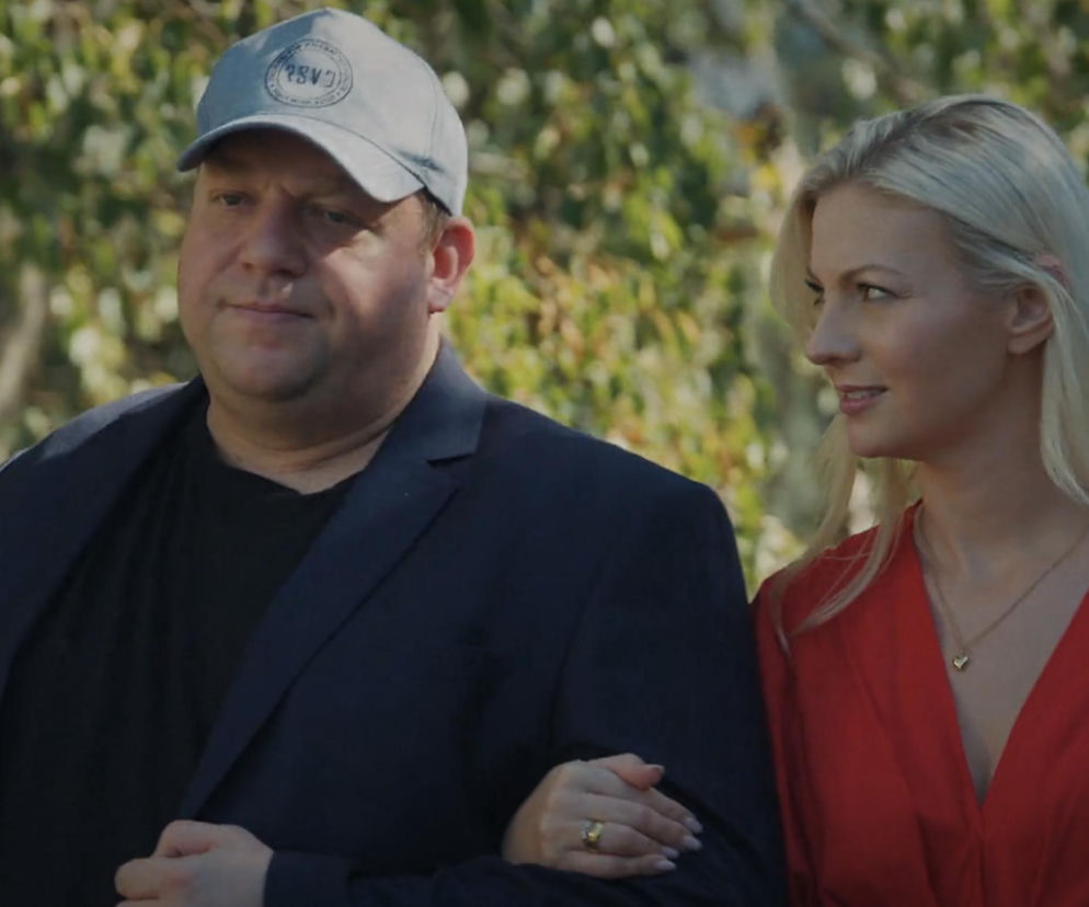 Tadeusz Kiemlicz w czapce z daszkiem w ciemnej marynarce i czarnym t-shircie, obok Jagoda w czerwonej sukience z naszyjnikiem w kształcie serca, która patrzy na niego z troską, trzymając go pod rękę. Scena z serialu M jak miłość dostępna na Super Seriale.