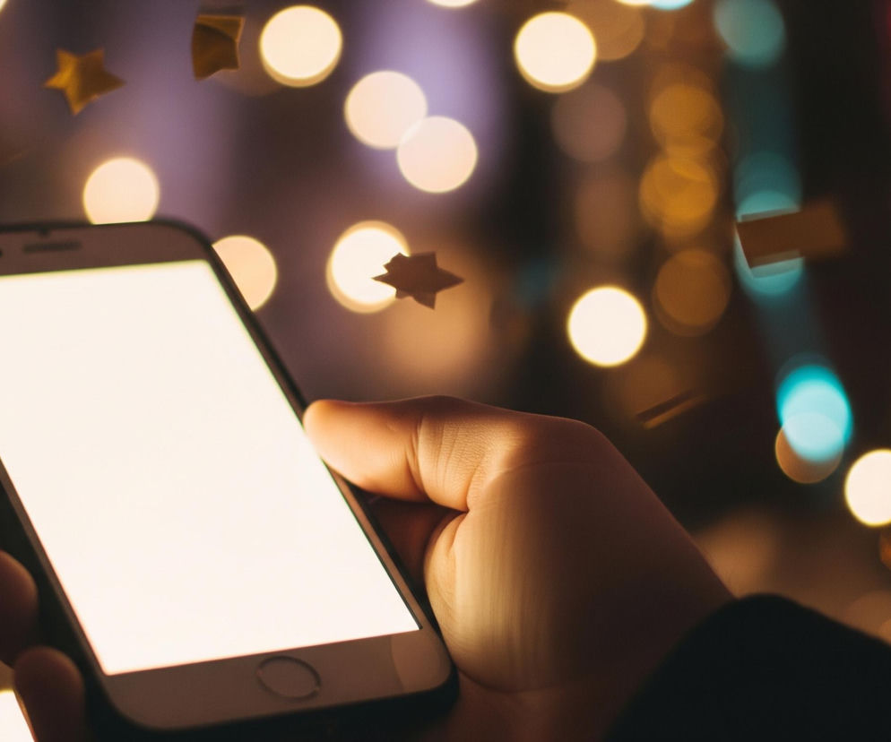 Prawa ręka trzyma smartfon z jasnym, białym ekranem skierowanym lekko w lewo. Kciuk spoczywa na górnej krawędzi urządzenia, a palce wskazujący i środkowy obejmują prawy bok, podczas gdy reszta dłoni podtrzymuje dolną część telefonu. Tło rozmyte, wypełniają je liczne okrągłe, jasne plamy światła w odcieniach ciepłego żółtego i pomarańczowego, a także kilka mniejszych, turkusowych i fioletowych akcentów. Pośrodku i w górnej części obrazu unoszą się złote elementy konfetti w kształcie gwiazdek i prostokątów.