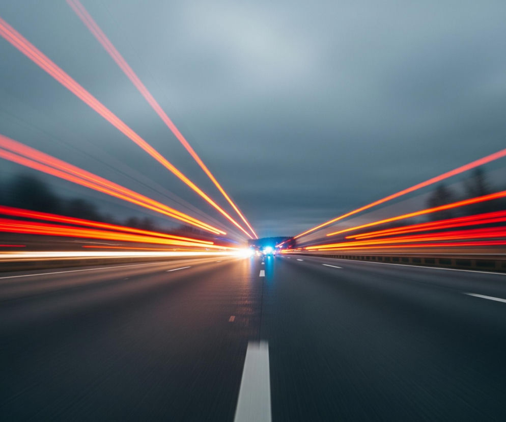 Czarna asfaltowa droga rozciąga się w głąb obrazu, podzielona na dwa pasy ruchu przerywaną białą linią centralną. Po obu stronach drogi widoczne są rozmazane czerwone i pomarańczowe smugi światła, biegnące równolegle do jezdni i zbiegające się w centralnym punkcie oddalonym. W tym punkcie zbiegu pojawiają się intensywne, jaskrawe światła, głównie niebieskie i białe, sugerujące jadące pojazdy. Niebo jest pochmurne, w odcieniach szarości i błękitu, z ciemnymi, rozmazanymi sylwetkami drzew na horyzoncie.