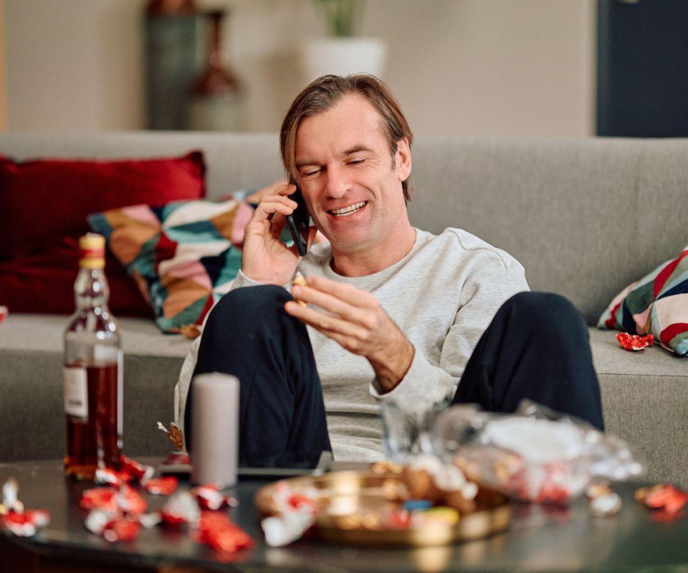 Mariusz (Piotr Cyrwus) w szarym swetrze i ciemnych spodniach, siedzi na podłodze, oparty o szarą kanapę, z uśmiechem rozmawia przez telefon. Na stoliku przed nim widoczna butelka alkoholu, słodycze i szklanka, co odzwierciedla jego problemy z nałogiem. Więcej o losach bohatera na Super Seriale.