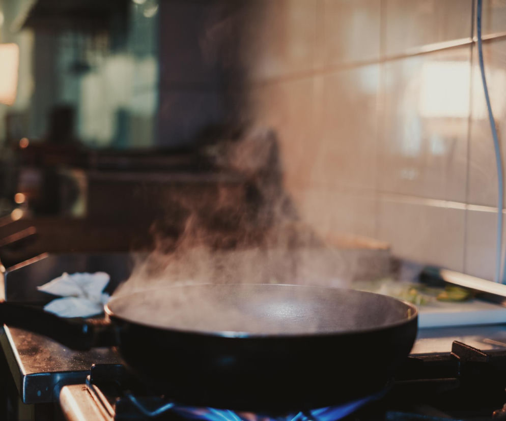 Patelnia na kuchence gazowej, z której unoszą się kłęby pary, symbolizujące szkodliwe opary ze smażenia, groźniejsze niż smog. Dowiedz się, jak skutecznie wietrzyć kuchnię i chronić swoje zdrowie, czytając artykuł na Poradnik Zdrowie.