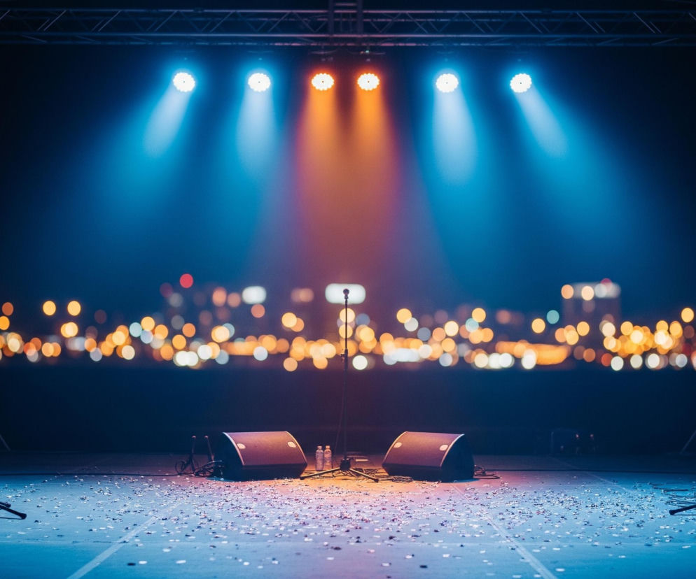 Na pierwszym planie widoczna jest scena, pokryta drobnymi kawałkami konfetti, częściowo oświetlona niebieskim światłem z lewej strony i pomarańczowym z prawej. Pośrodku sceny stoi mikrofon na statywie, otoczony dwoma czarnymi monitorami odsłuchowymi i dwoma butelkami wody. Po lewej stronie sceny opiera się gitara akustyczna, a obok niej leży pedalboard, natomiast po prawej stronie znajduje się zestaw perkusyjny. W tle, za sceną, rozpościera się rozmyty widok miasta z licznymi, ciepłymi, jasnymi punktami świetlnymi.