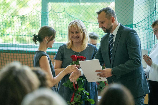 Mężczyzna w garniturze wręcza dokument nauczycielce z kwiatami. O szkoleniach dla pedagogów przeczytasz na SE Superbiz.