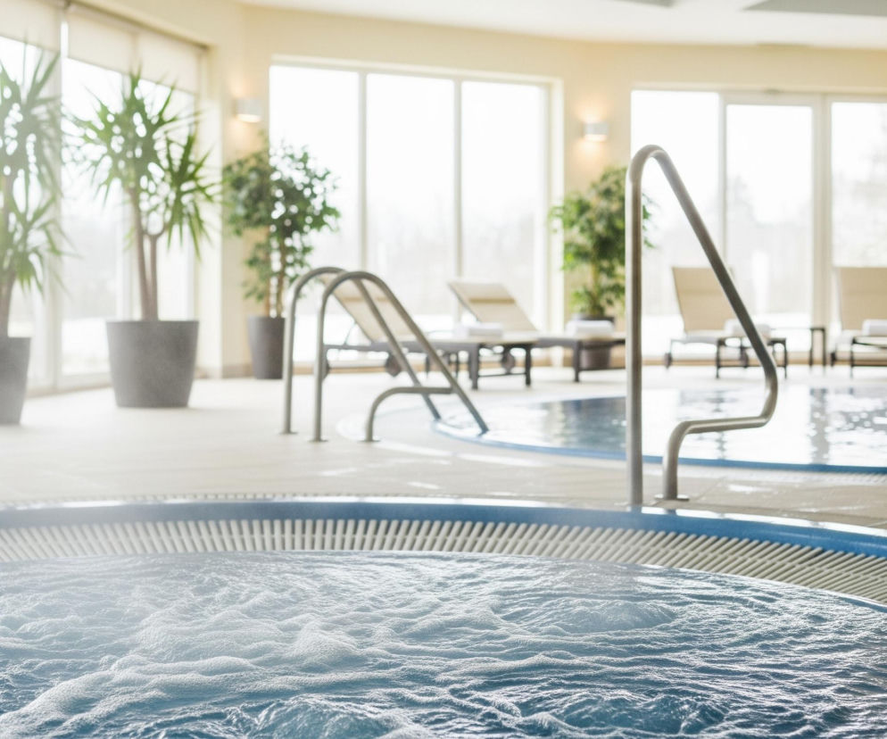 Na pierwszym planie, z lewej strony, widoczna jest powierzchnia wody w jacuzzi, pokryta drobnymi bąbelkami i delikatną mgiełką pary. Wokół błękitnego obrzeża jacuzzi znajduje się beżowa, antypoślizgowa kratka odpływowa. Dalej, w centrum obrazu, widać fragment basenu z metalowymi poręczami, a w tle rozciąga się przestronne pomieszczenie z dużymi oknami, przez które wpada jasne światło. W pomieszczeniu rozmieszczone są donice z wysokimi, zielonymi roślinami oraz leżaki.