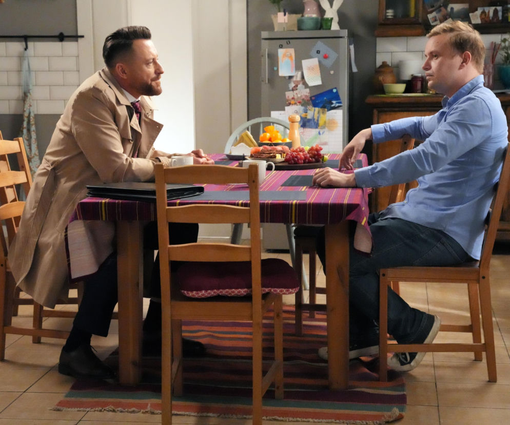 Marek Molak jako Hubert Pyrka, w jasnoniebieskiej koszuli i ciemnych jeansach, siedzi przy stole w kuchni i rozmawia z mężczyzną w beżowym płaszczu i garniturze. Na stole są owoce i kawa. Scena z serialu Barwy szczęścia, której opis dostępny jest na portalu Super Seriale.