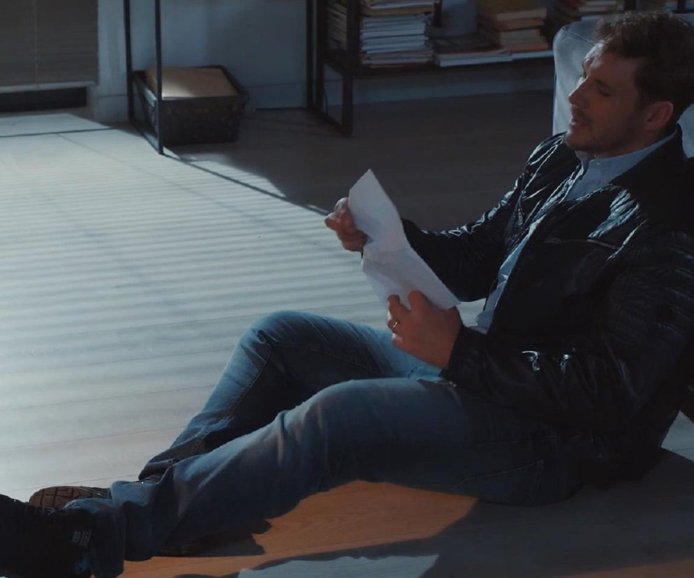 Mężczyzna w czarnej skórzanej kurtce i jeansach siedzący na podłodze, oparty o szarą kanapę, z pogiętą kartką papieru w dłoni, co symbolizuje jego rozpacz po odejściu Kamy w serialu M jak miłość. Więcej o tym odcinku przeczytasz na Super Seriale.