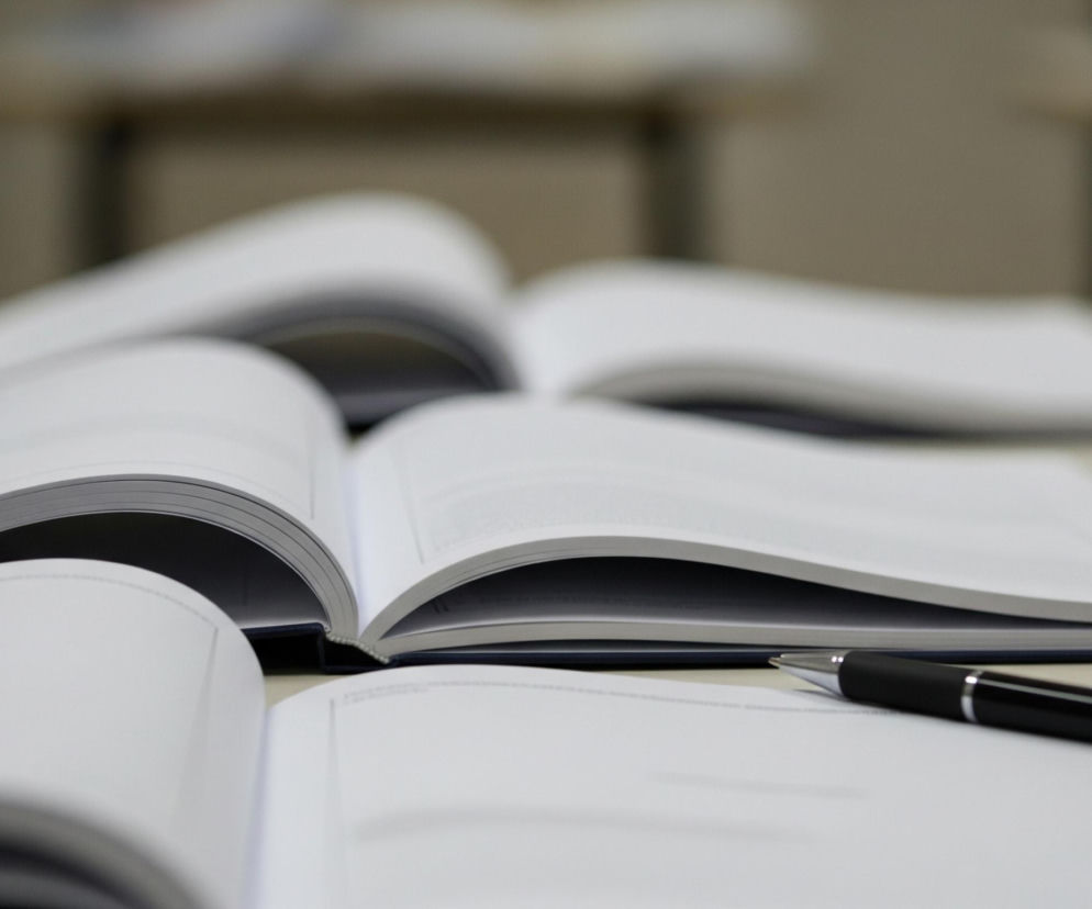 Na beżowym blacie leżą cztery otwarte książki z jasnymi stronami. W centrum kadru znajduje się pióro w kolorze czarnym z metalicznymi elementami, leżące na otwartej książce, której strony są głównie puste. Tło jest rozmyte, ukazując niewyraźne kształty innych obiektów i stolików.