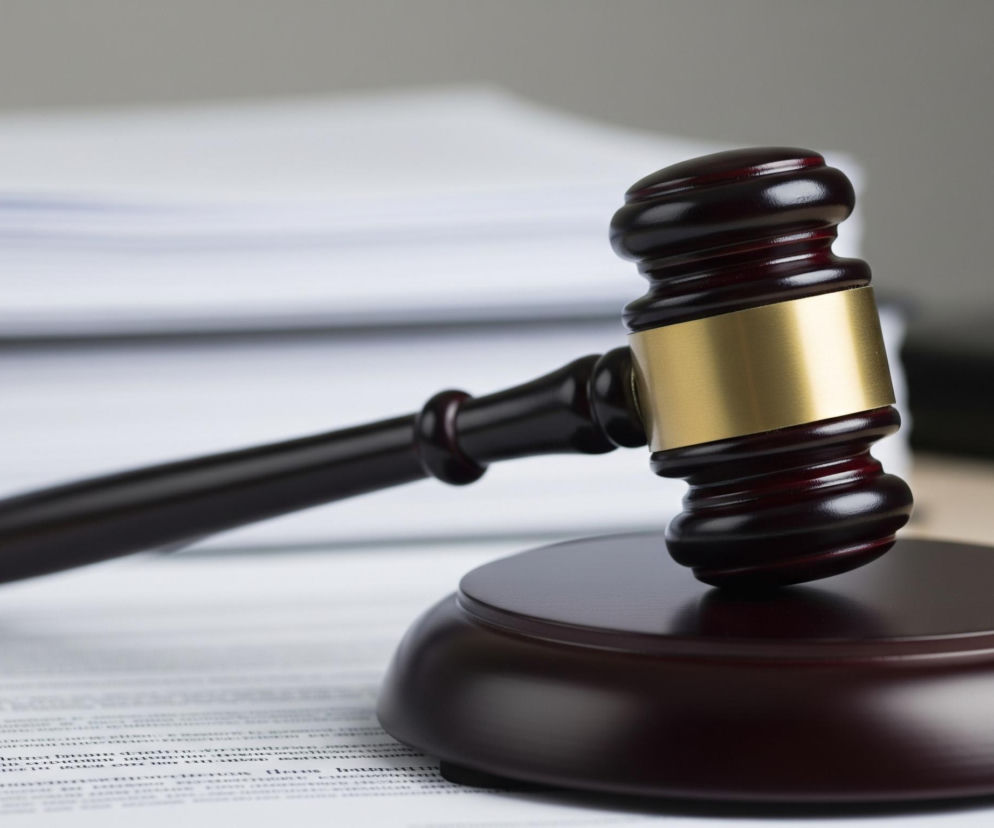 Drewniany młotek sędziowski w kolorze ciemnobrązowym spoczywa na dopasowanej podstawie, również ciemnobrązowej. Obuch młotka jest ozdobiony złotą opaską, a rączka leży poziomo na stosie białych kartek. Tło jest rozmyte, z widocznymi odcieniami szarości i bieli, co sugeruje biurowe otoczenie.