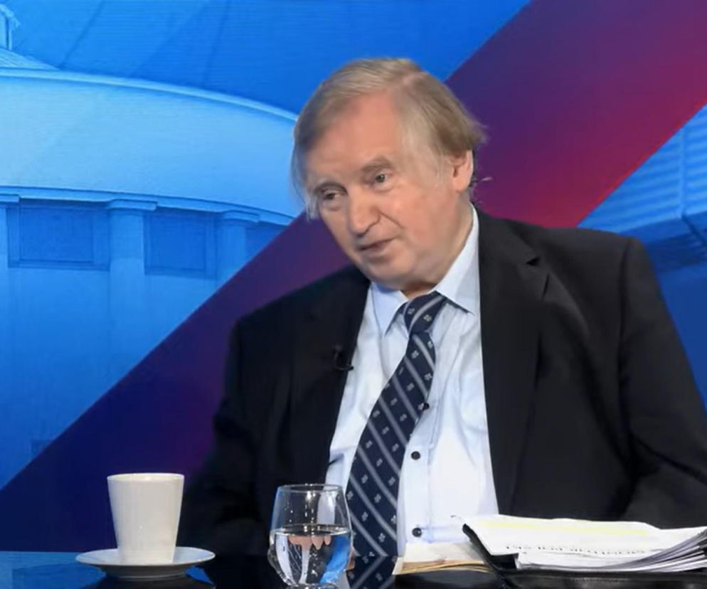 Profesor Ryszard Piotrowski, konstytucjonalista, w garniturze i krawacie, siedzi przy stole, obok filiżanka kawy i szklanka wody, w tle niebieska grafika z elementami architektonicznymi. Ekspert komentuje bieżące wydarzenia polityczne, o których więcej przeczytasz na naszym portalu.