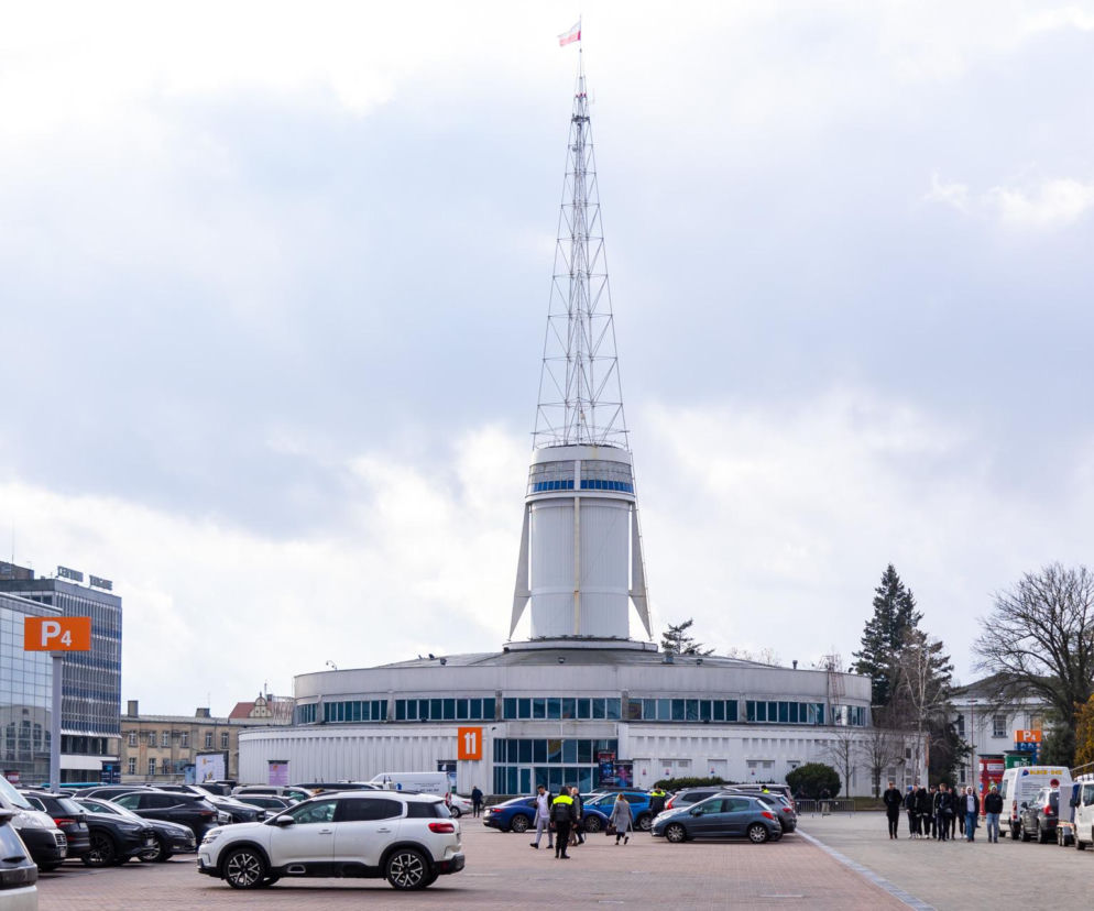 Budynek Iglicy Międzynarodowych Targów Poznańskich z polską flagą na szczycie, otoczony parkingiem z samochodami i ludźmi. Zdjęcie ilustruje kontekst artykułu o społecznikach walczących o nową PeWuKę w Poznaniu. Więcej na ten temat przeczytasz na naszym portalu.