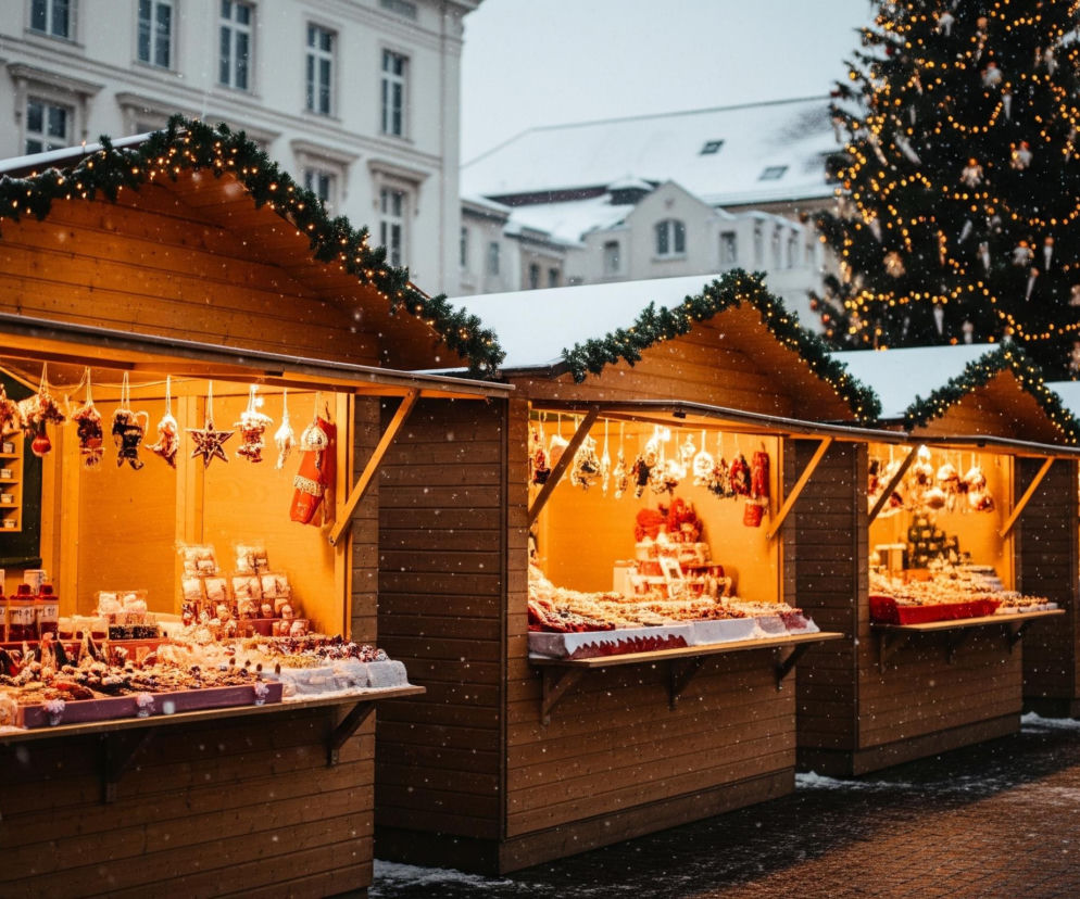 W centrum zdjęcia widoczny jest rząd drewnianych straganów świątecznych, oświetlonych ciepłym, pomarańczowym światłem, z zaśnieżonymi dachami udekorowanymi girlandami i małymi lampkami. Na blatach straganów, wyłożonych białymi i czerwonymi tkaninami, prezentowane są liczne słodycze, w tym ciastka i cukierki, a także suszone owoce i inne ozdoby. W tle, po prawej stronie, majestatycznie wznosi się duża, udekorowana choinka świąteczna z licznymi złotymi lampkami i bombkami, podczas gdy po lewej i w centrum widoczne są zaśnieżone budynki miejskie, z oknami w regularnych rzędach. Drobne płatki śniegu delikatnie opadają na całą scenę, pokrywając dachy i ulicę, tworząc zimową, świąteczną atmosferę.