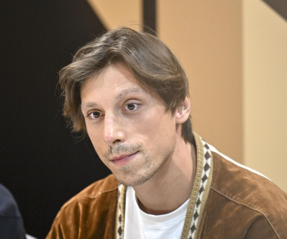 Aktor Eryk Kulm w brązowej marynarce i białym t-shircie spogląda w bok. O jego wielkiej metamorfozie przeczytasz na SE.