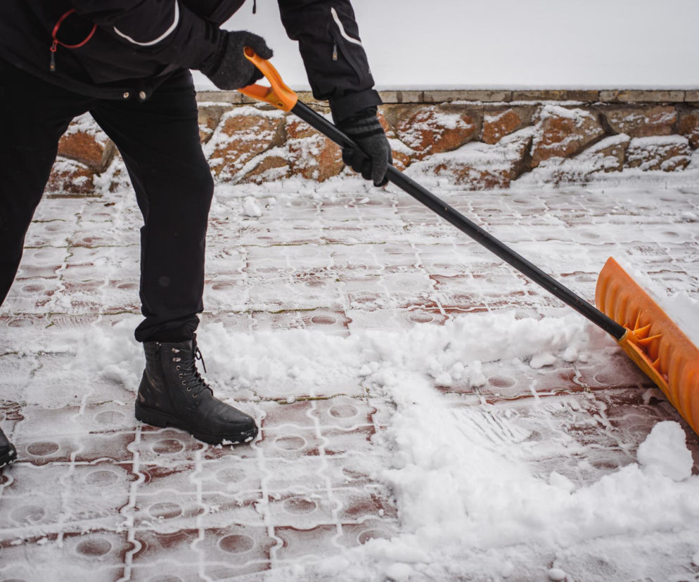 Osoba w czarnych butach i kurtce odśnieżająca chodnik przed domem pomarańczową łopatą. Mróz i intensywny wysiłek podczas odśnieżania mogą obciążyć serce, co może być szczególnie niebezpieczne dla osób z chorobami serca. Więcej na ten temat przeczytasz na Poradnik Zdrowie.