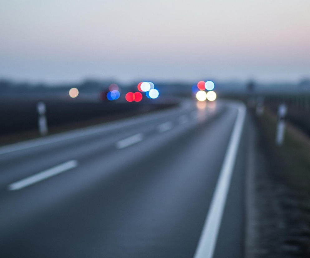 Asfaltowa droga biegnie ukośnie przez centrum kadru, rozszerzając się ku dołowi, z widocznymi przerywanymi białymi liniami. Po lewej stronie drogi rozciąga się ciemne, niewyraźne pole, a po prawej widoczny jest podobnie rozmyty, trawiasty teren z kilkoma słupkami. W oddali, na horyzoncie, znajdują się rozmazane światła pojazdów, głównie czerwone i niebieskie, oraz jaśniejsze, żółtawe światła z przodu. Niebo jest szare u góry i lekko różowofioletowe bliżej horyzontu.