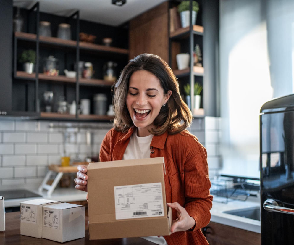 Uśmiechnięta kobieta w rudym ubraniu otwiera paczkę w kuchni, na blacie leżą inne przesyłki; sprawdź na Poradnik Zdrowie, dlaczego produkty z Temu i Shein są często niebezpieczne, zawierają toksyny, a zabawki grożą uduszeniem.