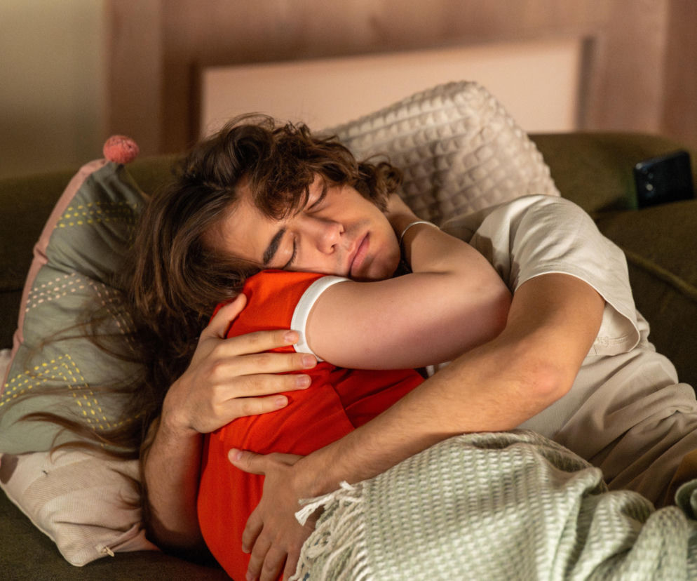 Młody mężczyzna, Romeo, śpi na zielonej kanapie, obejmując poduszkę w czerwonej poszewce. Ma na sobie beżową koszulkę i jest przykryty jasnym, wzorzystym kocem. Obok na poduszce widać leżący telefon. Scena odzwierciedla miłosny zgrzyt u Grzelaków z serialu Barwy szczęścia, o czym więcej przeczytasz na Super Seriale.