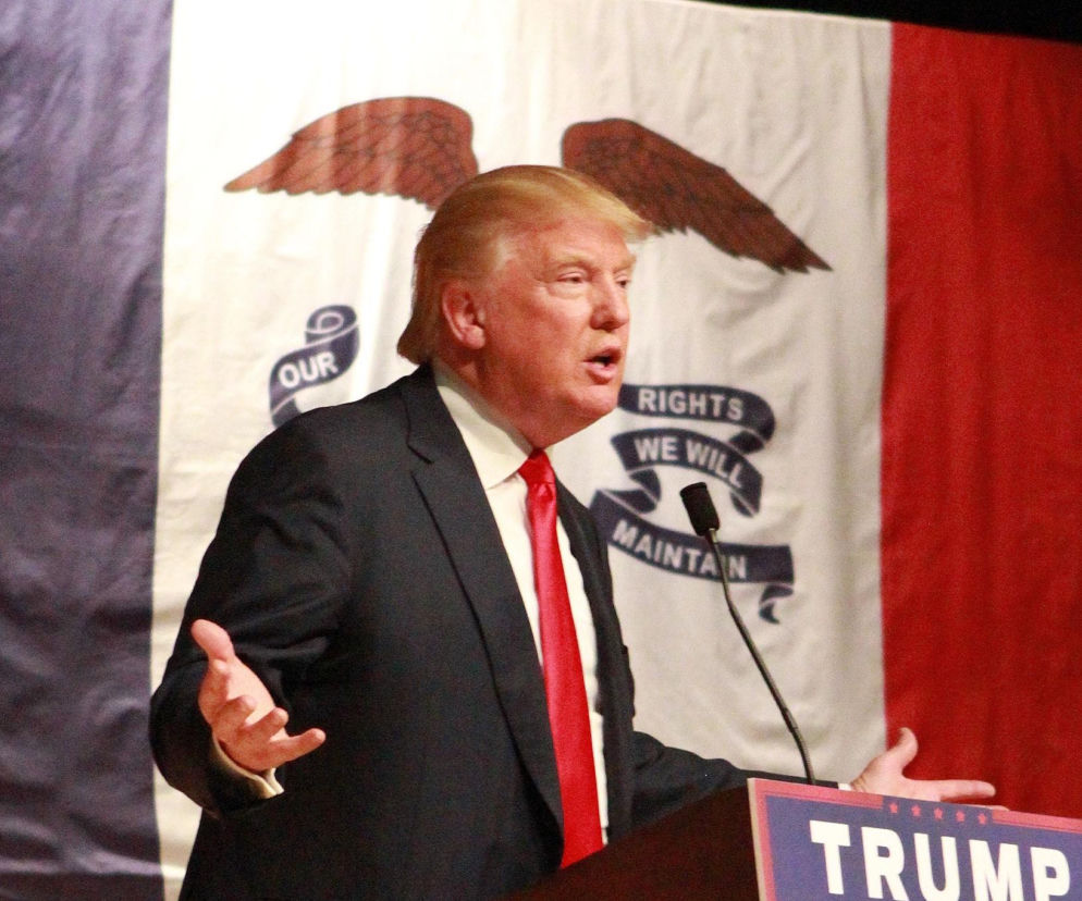 Donald Trump przemawia za pulpitem na tle flagi stanu Iowa. O sondażach poparcia przeczytasz na SE Portal Obronny.