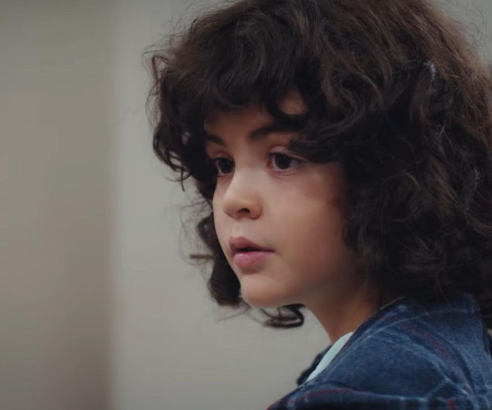 Chłopiec Yusuf z serialu Dziedzictwo, o ciemnych kręconych włosach i ciemnych oczach, ubrany w koszulę w kratę, spogląda z zatroskaniem. Jego twarz wyraża niepokój, co oddaje emocje związane z fabułą serialu. O najnowszych odcinkach dowiesz się na Super Seriale.