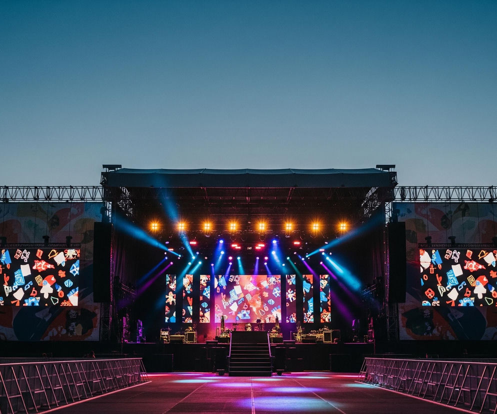 Rozświetlona scena festiwalowa dominuje centralnie, z widocznymi konstrukcjami kratownicowymi po bokach i na górze, wspierającymi ekrany LED i reflektory. Trzy duże ekrany LED wyświetlają kolorowe, abstrakcyjne wzory, a wiele reflektorów na górze sceny emituje jaskrawe, niebieskie, fioletowe i bursztynowe światła. Przed sceną rozciąga się pusta przestrzeń z rzędami metalowych barierek, oświetlona kolorowymi światłami odbijającymi się od podłoża. Tło stanowi stopniowo ciemniejące niebo, przechodzące od jasnego błękitu u góry do cieplejszego odcienia pomarańczu i różu na horyzoncie, z zarysami drzew i namiotów po lewej stronie.