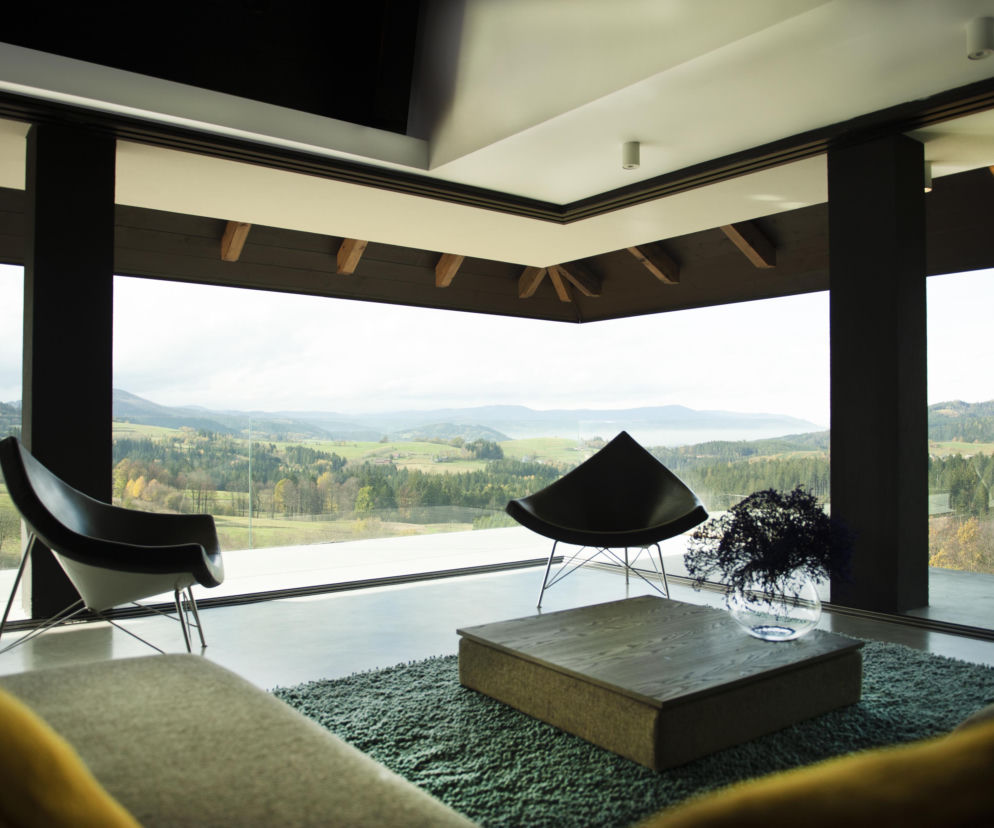 Nowoczesny salon w Veranda Villa w Istebnej z panoramicznymi oknami, otwierający widok na malowniczy krajobraz Beskidu Śląskiego. Dwie designerskie fotele, niski stolik kawowy na zielonym dywanie, betonowa podłoga, sufit z drewnianymi krokwiami. Architektura Murator Plus.