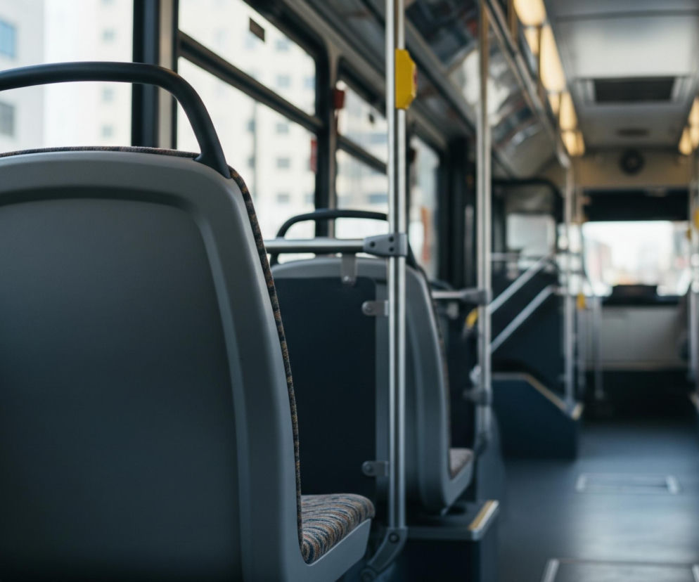 Wnętrze pustego autobusu miejskiego widoczne jest z perspektywy siedzenia, skupiając się na oparciu przedniego fotela w kolorze szarym. Po prawej stronie rozciąga się rząd siedzeń z tapicerką w brązowo-granatowo-beżowe pasy, przymocowanych do metalowych poręczy biegnących przez całą długość pojazdu. Przez duże okna z lewej strony widać fragmenty jasnych budynków z prostokątnymi oknami, rozmytych w tle, natomiast wnętrze autobusu oświetlają podłużne lampy sufitowe.