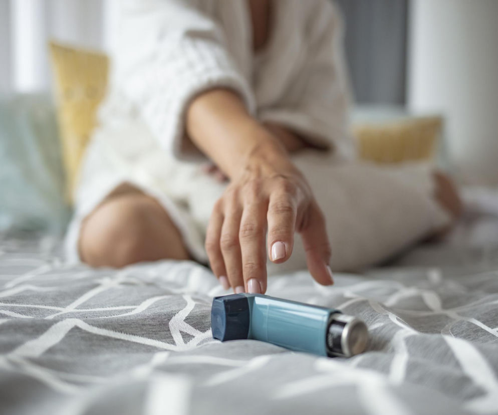 Kobieta w białym szlafroku na łóżku, z ręką wyciągniętą po inhalator, symbolizujący walkę z dusznością i astmą. Dowiedz się, jak nowe terapie mogą przywrócić normalność w walce z ciężką astmą, czytając na Poradnik Zdrowie.