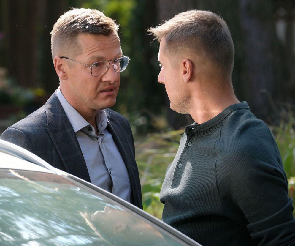 Paweł i Piotrek Zduńscy z M jak miłość rozmawiają przy samochodzie. Paweł w okularach i marynarce, Piotrek w koszulce polo, stoją blisko siebie, patrząc na siebie. Scena pożegnania braci, pełna emocji, dostępna na Super Seriale.