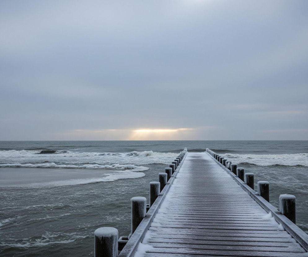 Drewniane molo, pokryte cienką warstwą śniegu, rozciąga się w głąb wzburzonego, ciemnoszarego morza, zajmując środek dolnej części kadru i prowadząc wzrok ku horyzontowi. Po obu stronach mola widać spienione fale, uderzające o piaszczysty brzeg po lewej stronie i tworzące białe pasma na powierzchni wody. Nad horyzontem, w centrum obrazu, wąska, pozioma linia jaśniejszego, żółtego światła przebija się przez ciężkie, szare chmury, rzucając delikatne promienie w dół.