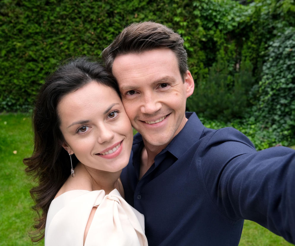 Kasia (Paulina Lasota) i Jakub (Krzysztof Kwiatkowski) z M jak miłość uśmiechają się do selfie, stojąc blisko siebie na tle zielonych krzewów i trawy. Paulina ma na sobie jasną, elegancką sukienkę, a Krzysztof ciemną koszulę. Więcej o ich losach w Super Seriale.