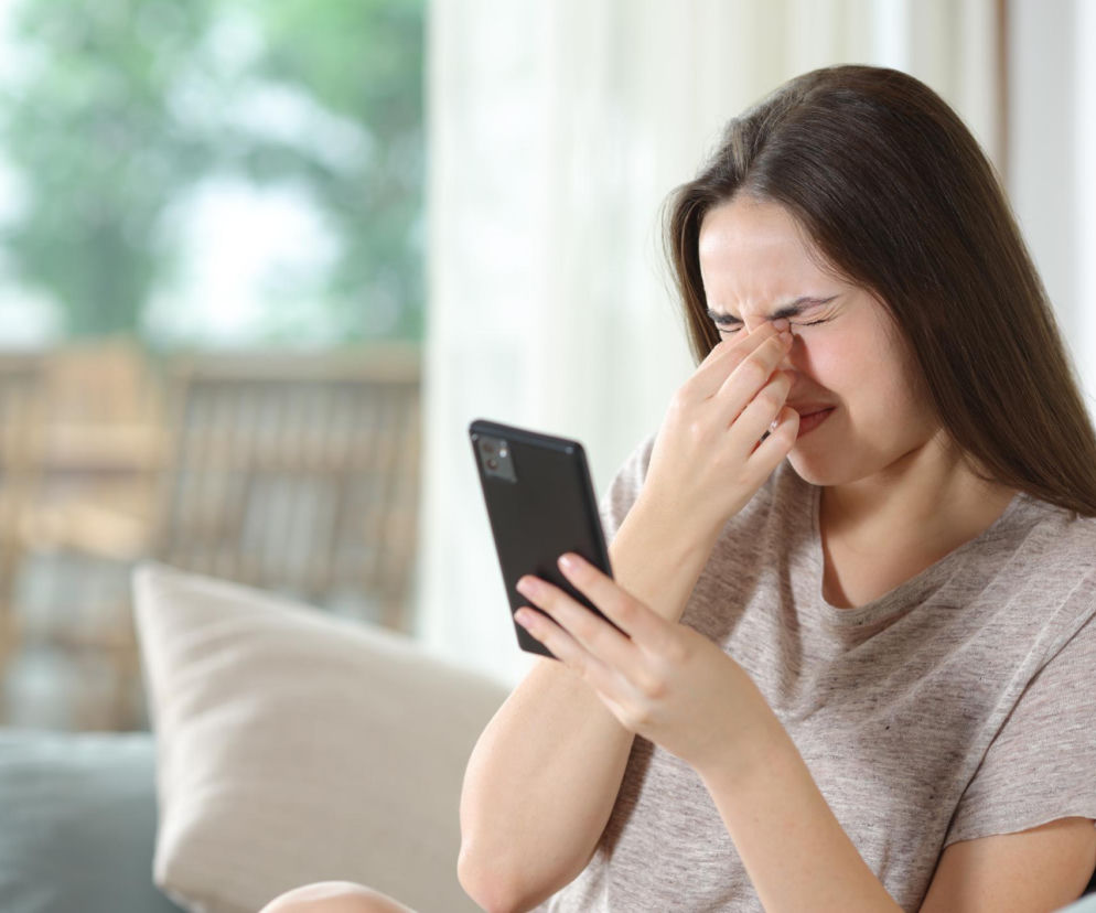 Kobieta z zamkniętymi oczami, masująca skronie i trzymająca w dłoni smartfon. Odczuwa ból głowy lub mdłości od telefonu, co jest objawem cyfrowej choroby lokomocyjnej. O jej przyczynach i metodach zapobiegania przeczytasz na portalu Poradnik Zdrowie.