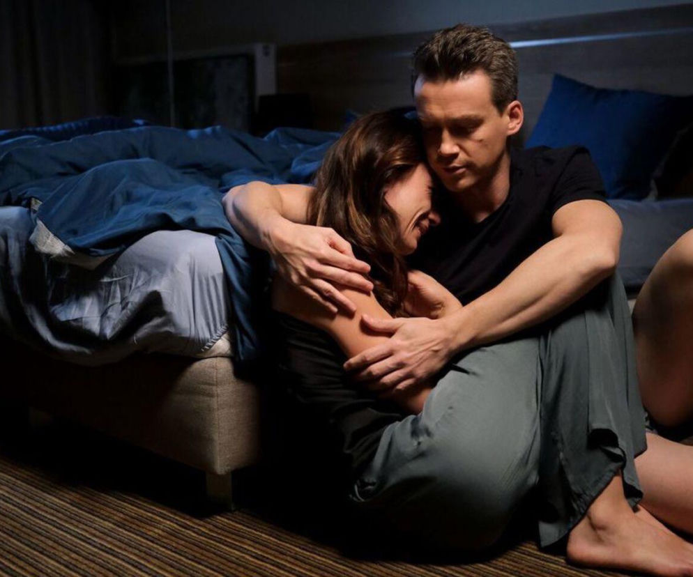 Martyna (Magdalena Turczeniewicz) i Jakub (Krzysztof Kwiatkowski) z serialu M jak miłość siedzą na podłodze w hotelowym pokoju. Jakub obejmuje płaczącą Martynę, która tuli się do niego w geście pocieszenia, po zobaczeniu zdjęć ślubnych Marcina. O tych emocjonalnych scenach przeczytasz na stronie Super Seriale.