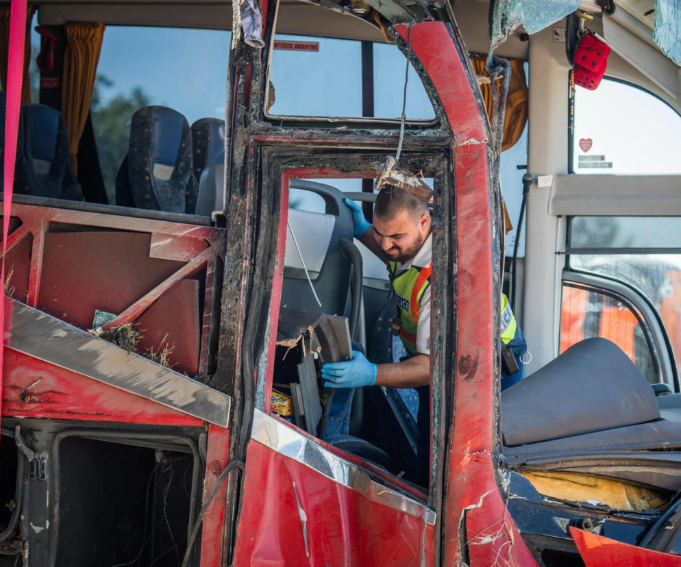 Mężczyzna w niebieskich rękawiczkach i kamizelce przy rozbitym czerwonym autokarze. O katastrofie na Węgrzech pisze SE.