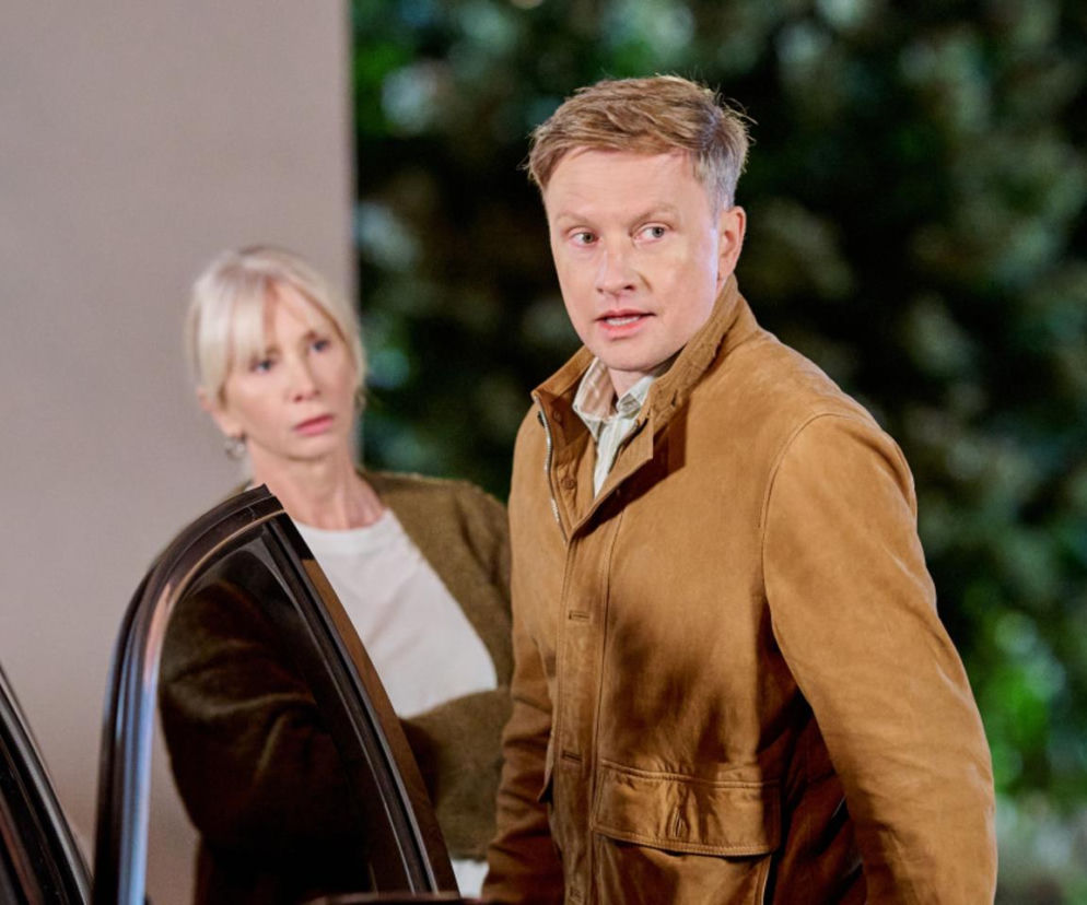 Aktorzy Ewa Gawryluk jako Ewa i Jakub Wesołowski jako Igor z serialu Na Wspólnej, stoją obok otwartych drzwi samochodu. Igor patrzy w lewo z otwartymi ustami, wyrażając zdziwienie lub irytację. Ewa patrzy w prawo z troską. Więcej o ich losach na Super Seriale.