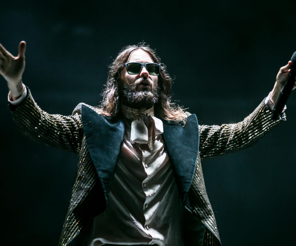 Muzyk Jared Leto w ciemnych okularach i błyszczącej marynarce trzyma mikrofon. O zarzutach wobec gwiazdy przeczytasz w Eska.