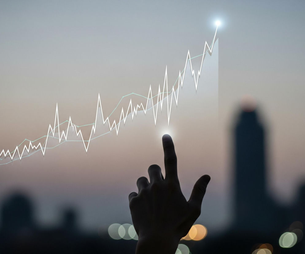 Na pierwszym planie widoczna jest ciemna sylwetka dłoni, z palcem wskazującym dotykającym wirtualnej linii wykresu, z której emanuje jasne, białe światło. Wykres składa się z dwóch linii: grubszej, nieregularnej, białej linii o ostrych szczytach i dolinach, oraz cieńszej, jasnozielonej linii podążającej za ogólnym trendem wzrostowym. Obie linie wznoszą się dynamicznie w prawą stronę, a biała linia osiąga szczyt zakończony intensywnym, białym blaskiem. Tło stanowi rozmyta panorama miasta o zmierzchu, z ciemnymi zarysami budynków i rozświetlonymi, rozmazanymi światłami w dolnej części, a niebo przybiera odcienie szarości i błękitu przechodzące w delikatny róż.