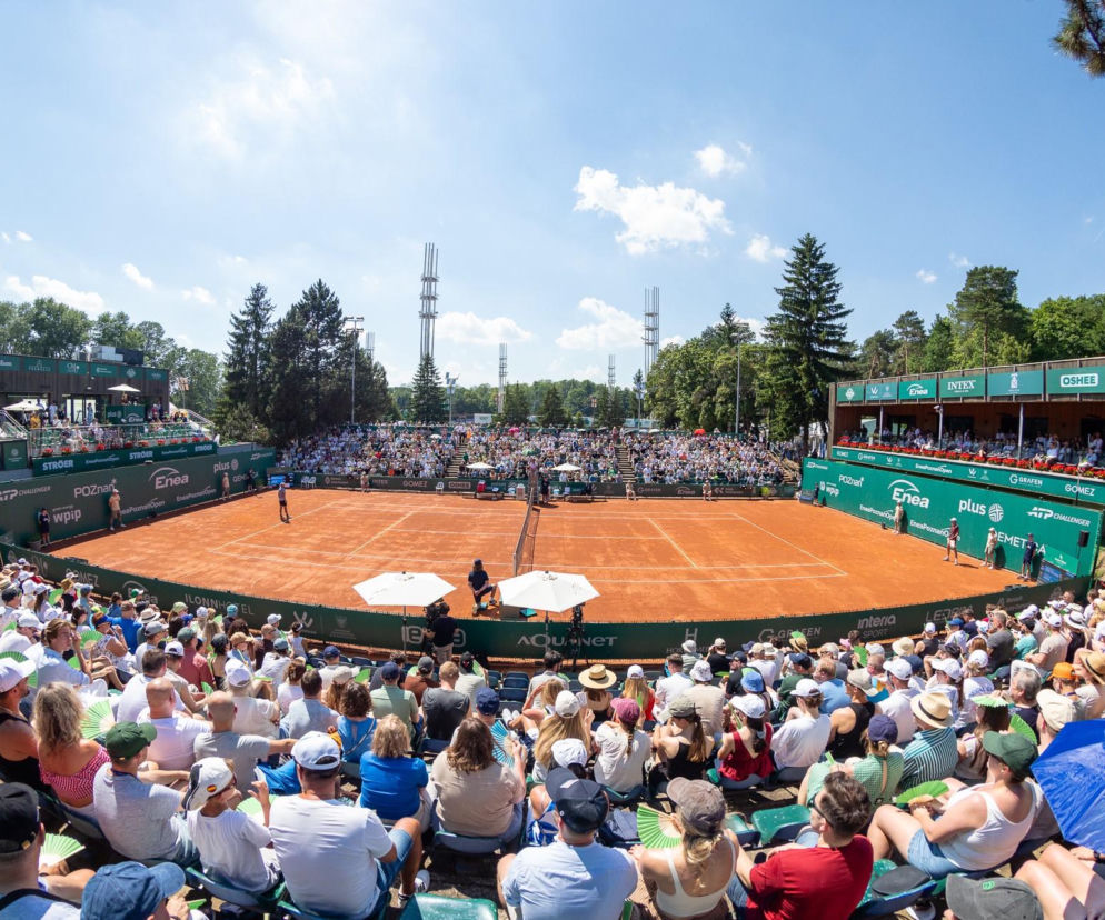 Widok z góry na kort tenisowy Enea Poznań Open, gdzie trwa mecz, a trybuny wypełnia tłum kibiców, wielu z nich z wachlarzami i parasolami, widoczne są bannery sponsorów. O wydarzeniu przeczytasz więcej na naszym portalu.