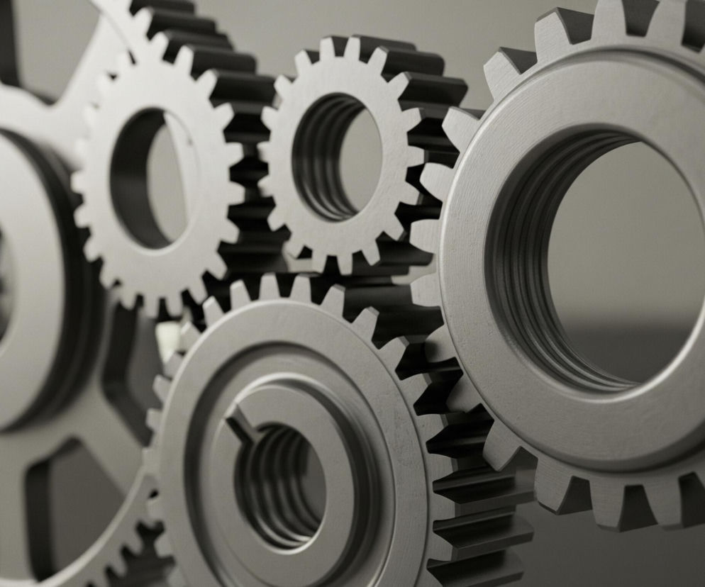 Na obrazie widoczne są mechanizmy zębate w różnych rozmiarach i głębi ostrości, utrzymane w monochromatycznej palecie szarości. Duże koło zębate po lewej stronie posiada wycięcia w ramionach, a mniejsze koła zazębiają się ze sobą, tworząc złożony układ. Detale powierzchni kół, w tym delikatne zarysowania i metaliczny połysk, są wyraźnie widoczne, szczególnie na pierwszym planie.