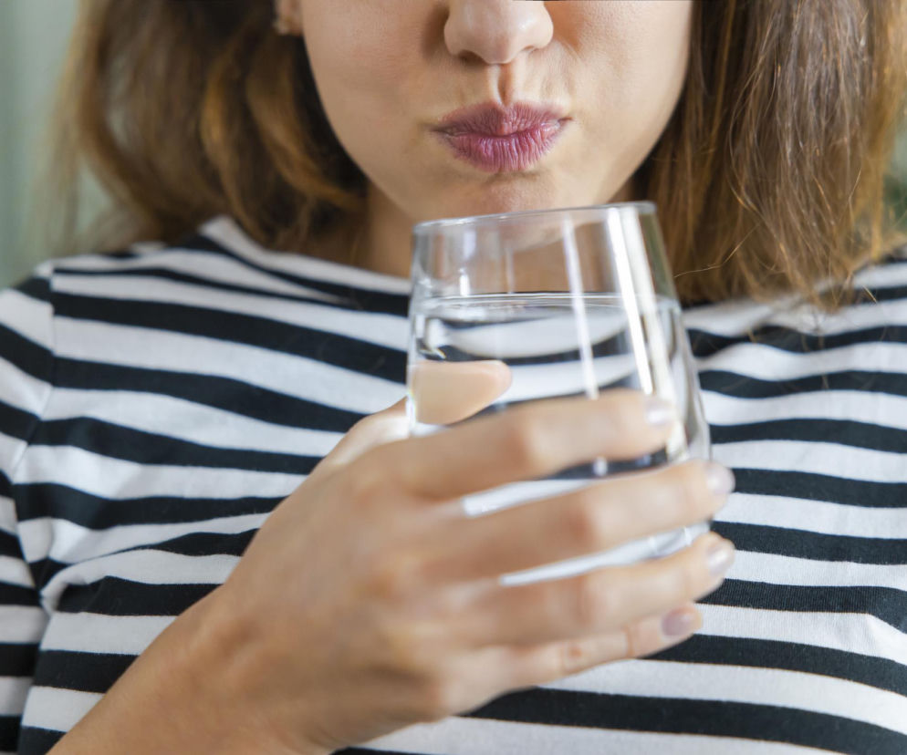 Kobieta płucze usta wodą po myciu zębów, trzymając szklankę. Ten częsty błąd niweczy ochronę przed próchnicą. Sprawdź na Poradnik Zdrowie, dlaczego płukanie ust po szczotkowaniu osłabia działanie fluoru i jak prawidłowo dbać o higienę jamy ustnej.