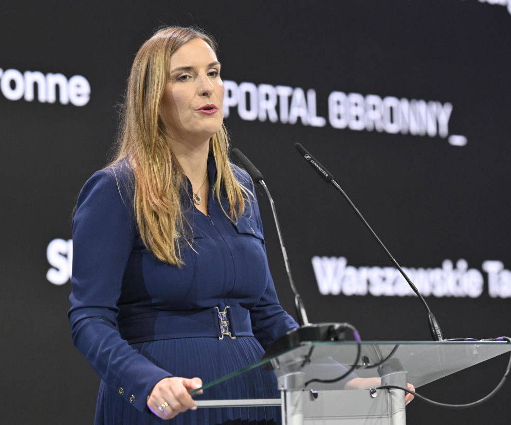Magdalena Sobkowiak-Czarnecka w granatowej sukience z paskiem, przemawiająca zza przezroczystej mównicy z mikrofonami Sennheiser podczas Warszawskich Targów Obronnych Expo XXI. Tło stanowi czarna ściana z białym napisem PORTAL OBRONNY, o zmianach roli pani minister czytaj na naszym portalu.