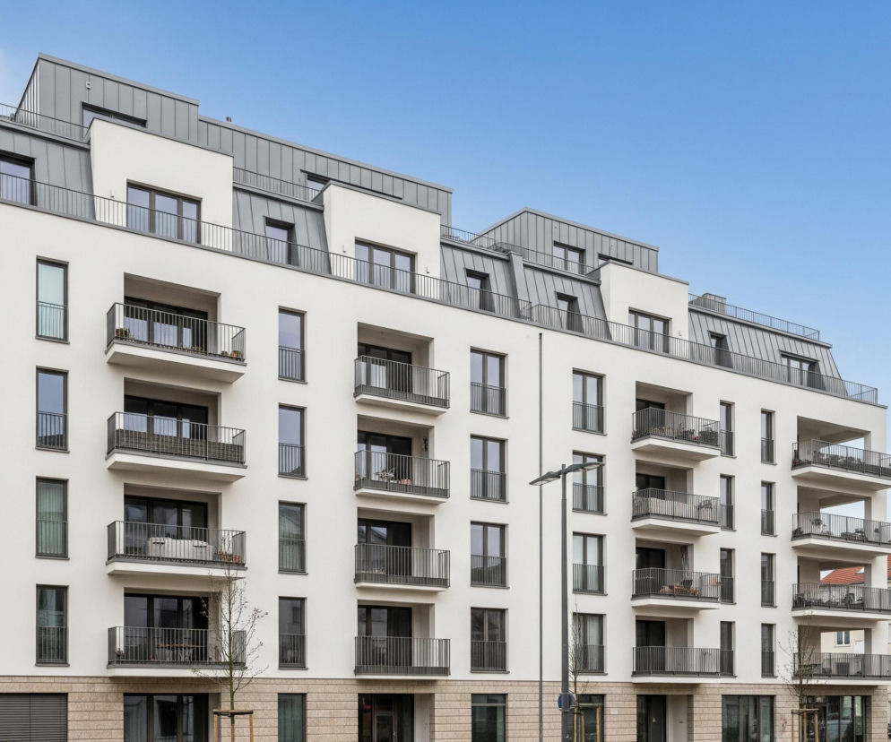 Nowoczesny budynek mieszkalny o sześciu kondygnacjach dominuje na zdjęciu, z białymi fasadami i ciemnoszarymi oknami oraz balustradami balkonów. Konstrukcja posiada zróżnicowany, skośny dach pokryty szarymi, metalowymi panelami, a na parterze widoczne są fragmenty elewacji z beżowego kamienia. Niebieskie, bezchmurne niebo stanowi tło dla tego architektonicznego obiektu. Po prawej stronie widać fragmenty innych budynków mieszkalnych oraz ulicę z zaparkowanymi samochodami i słupem oświetleniowym.