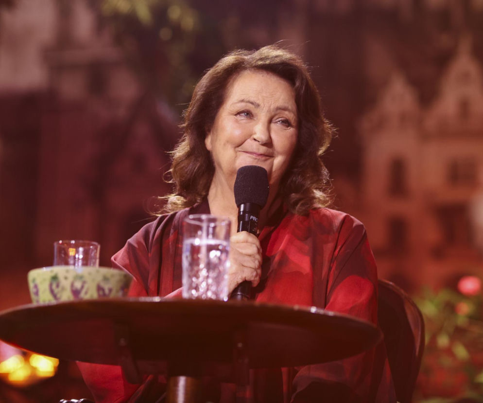 Uśmiechnięta Anna Dymna z mikrofonem siedzi przy stoliku. O metamorfozie gwiazdy filmu Znachor przeczytasz na SE.