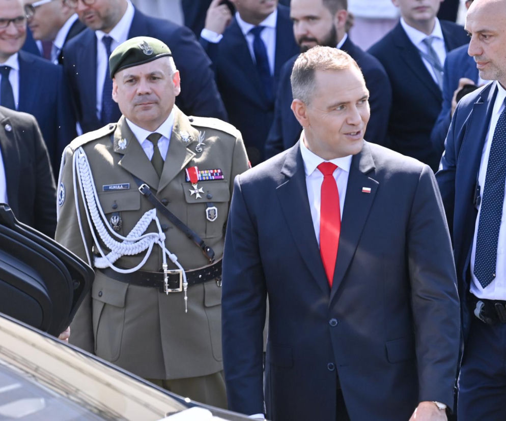 Karol Nawrocki w garniturze i czerwonym krawacie. Obok wojskowy i ochroniarze. O jego prezydenturze czytaj na SE Polityka.