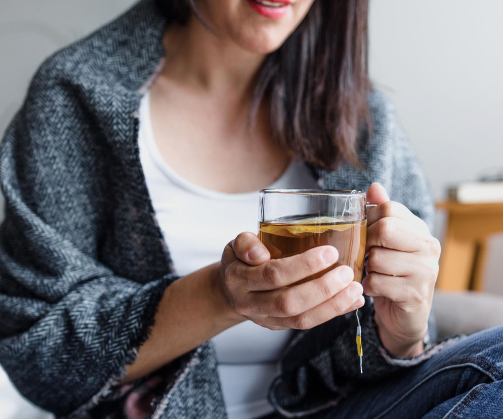 Kobieta w swetrze trzyma szklankę z ziołowym naparem, odczuwając ulgę po posiłku. Zdjęcie ilustruje domowe sposoby na wzdęcia i gazy, takie jak mięta czy koper włoski, o których szerzej przeczytasz na portalu Poradnik Zdrowie.
