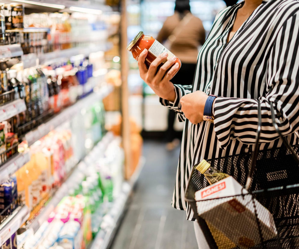 Kobieta w czarno-białej koszuli trzyma słoik sosu w supermarkecie, analizując etykietę produktu, co nawiązuje do tematu szkodliwych konserwantów w żywności. Więcej o wpływie dodatków na zdrowie przeczytasz na naszym portalu.