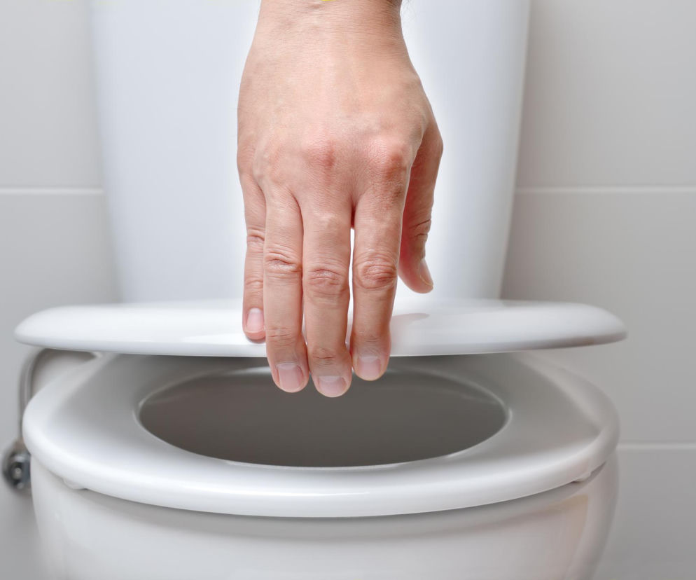 Ręka osoby podnoszącej deskę sedesową w czystej, białej toalecie, co symbolizuje ostrożność w korzystaniu z publicznych łazienek. Ta ilustracja nawiązuje do problemu unikania siadania na desce, który, jak informuje Poradnik Zdrowie, może prowadzić do infekcji pęcherza.