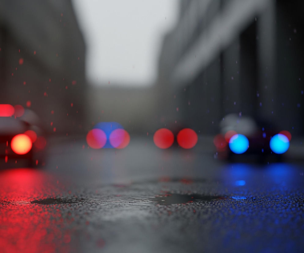 Mokra asfaltowa ulica rozciąga się w głąb obrazu, ukazując odległe samochody. Najbliższy, po lewej stronie, świeci czerwonymi światłami tylnymi, tworząc wyraźne odbicie na mokrej nawierzchni i delikatne, czerwone punkty, imitujące krople deszczu, unoszące się w powietrzu. Dalej, po prawej, widoczne są niebieskie światła samochodu, również odbijające się w kałużach i towarzyszące im niebieskie punkty. Ulica otoczona jest niewyraźnymi, ciemnymi budynkami, zdominowana przez szarości i czerń.