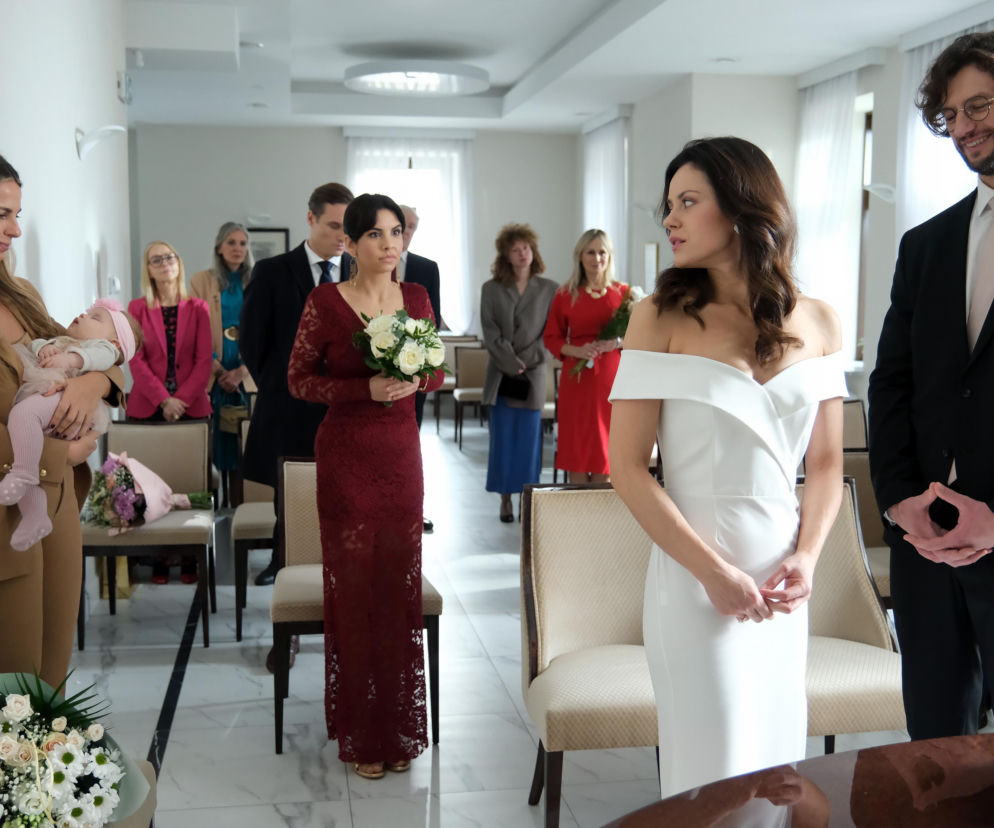 Paulina Lasota (Kasia) w białej sukni ślubnej i Mateusz Mosiewicz (Mariusz) w garniturze stoją przed ołtarzem, otoczeni gośćmi, w tym kobietą trzymającą dziecko i inną kobietą w bordowej sukience. Scena ze ślubu Kasi i Mariusza z M jak miłość. Więcej informacji o serialu znajdziesz na Super Seriale.